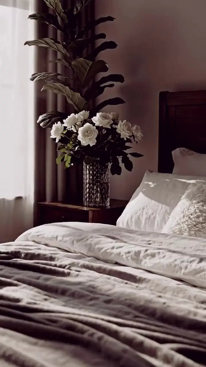 Quarto com a cama arrumada em roupa de cama clara, uma planta e a luz da janela